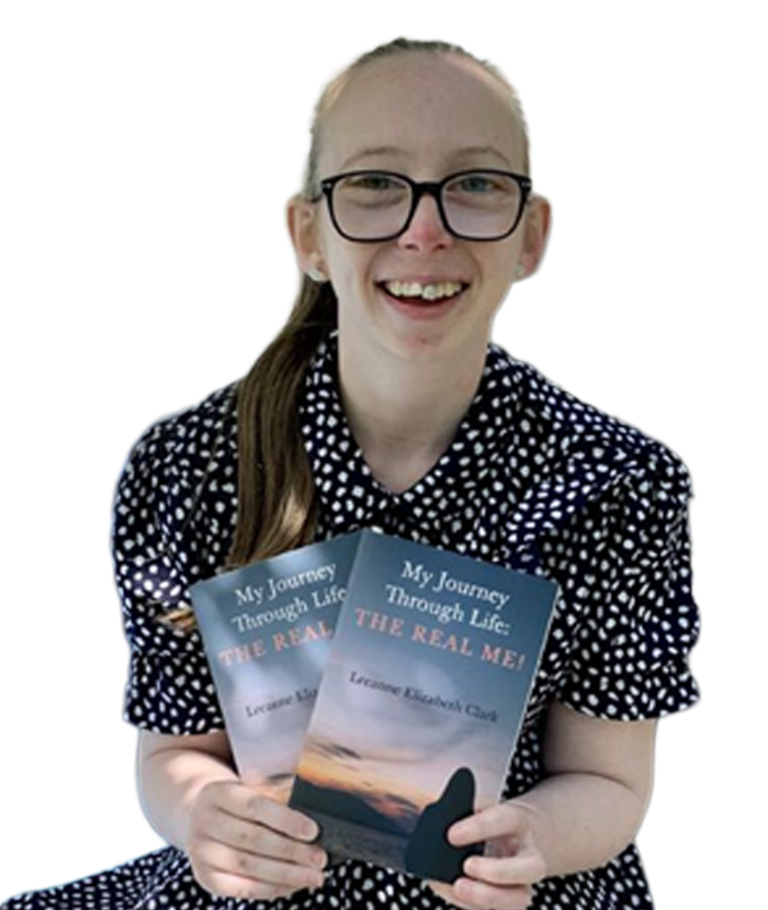 Leanne C, wearing glasses and a black polka dot shirt, smiling while holding copies of her book 'My Journey Through Life: The Real Me