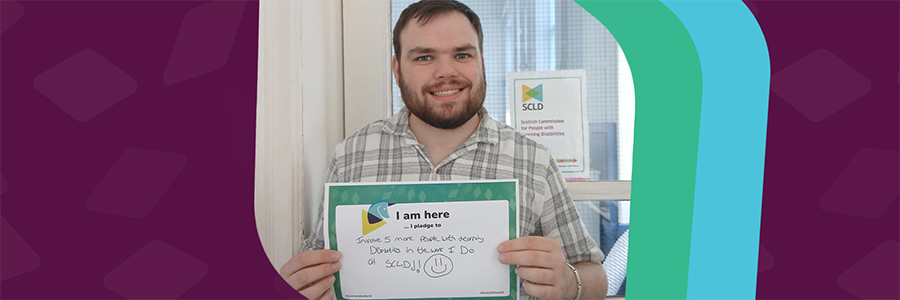 A man with short dark hair and a beard, wearing a plaid shirt, is smiling and holding a pledge sign in front of a door with an SCLD (Scottish Commission for People with Learning Disabilities) logo in the background. His pledge says I am here ... I pledge to involve 5 more people with learning disabilities in the work I do at SCLD!!