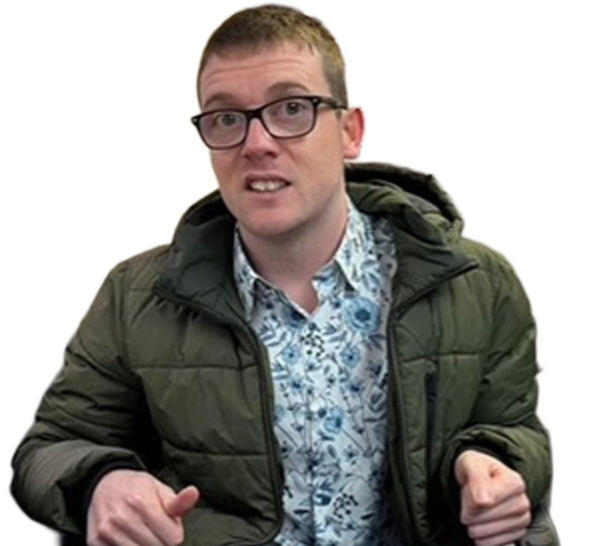 Sandy, wearing glasses and a green puffer jacket over a patterned shirt, gesturing while speaking